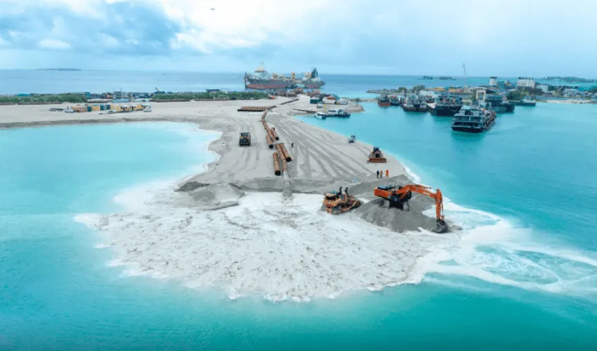 Excavators shaping a strip of newly reclaimed sand at Ras Malé, with dredging pipes and work boats in the lagoon behind