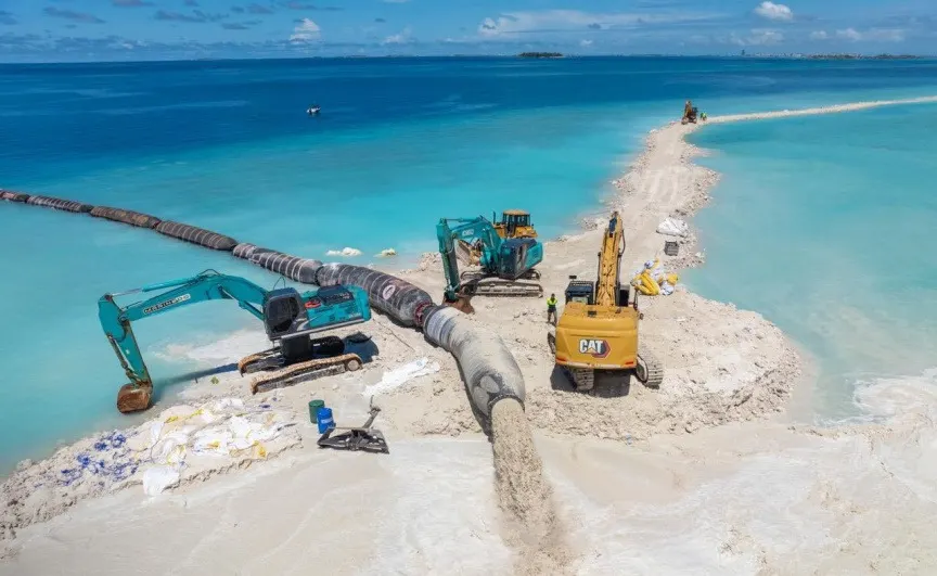 Excavators and a bulldozer working beside a large dredging pipeline on a narrow strip of reclaimed sand at Ras Malé