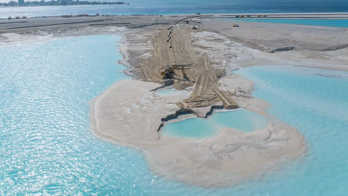Newly reclaimed sand being shaped by machinery in a turquoise lagoon, with a city skyline on the horizon