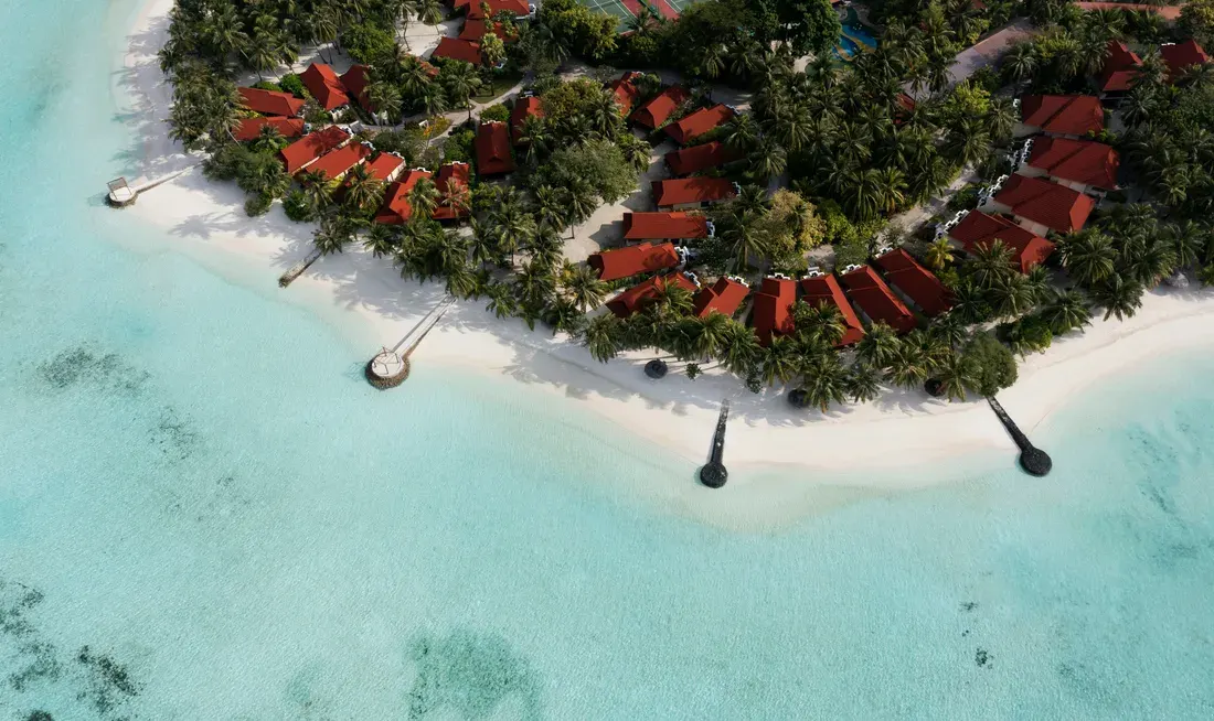 Aerial view of a resort island near Malé with a swimming pool, beach villas among palm trees and turquoise water