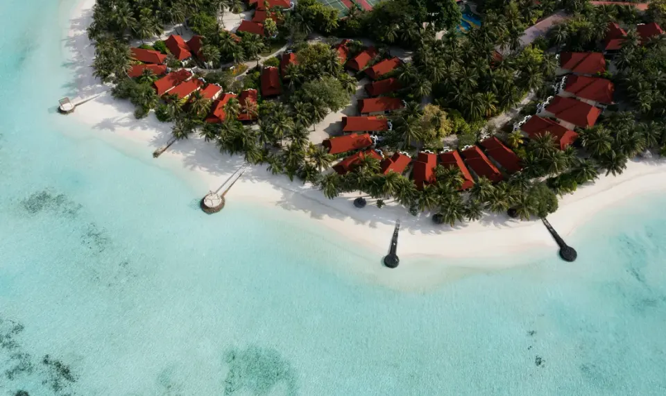 Aerial view of a resort island near Malé with a swimming pool, beach villas among palm trees and turquoise water