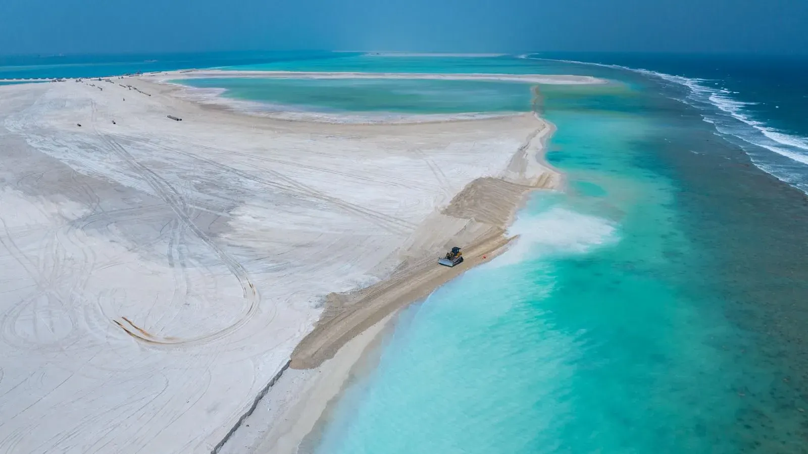 Aerial view of a wide stretch of newly reclaimed white sand at Ras Malé, with shallow turquoise water and the reef edge beyond